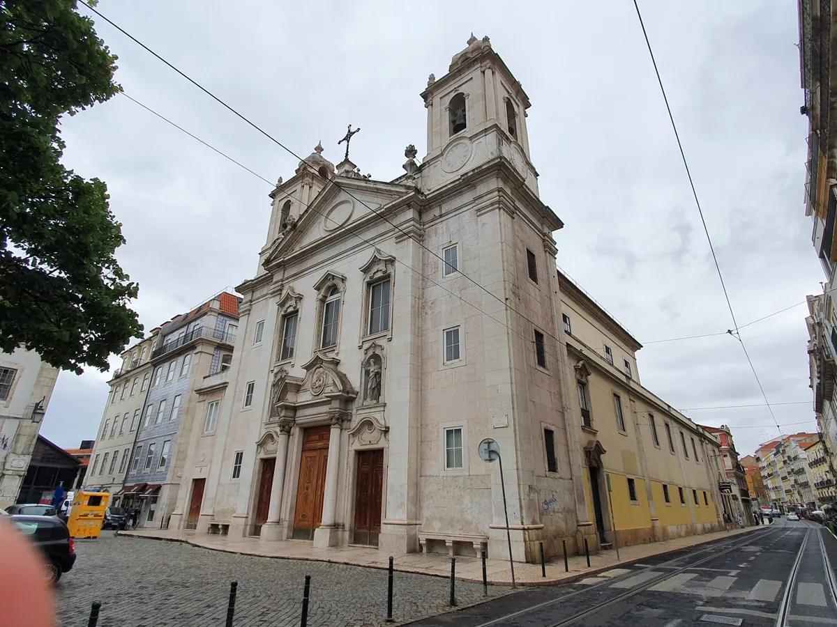 Kostel svatého Pavla v Lisabonu pohledem ze strany jeho náměstí pod zataženou oblohou, zvonice a trojúhelníkový štít nad trojicí dřevěných dveří